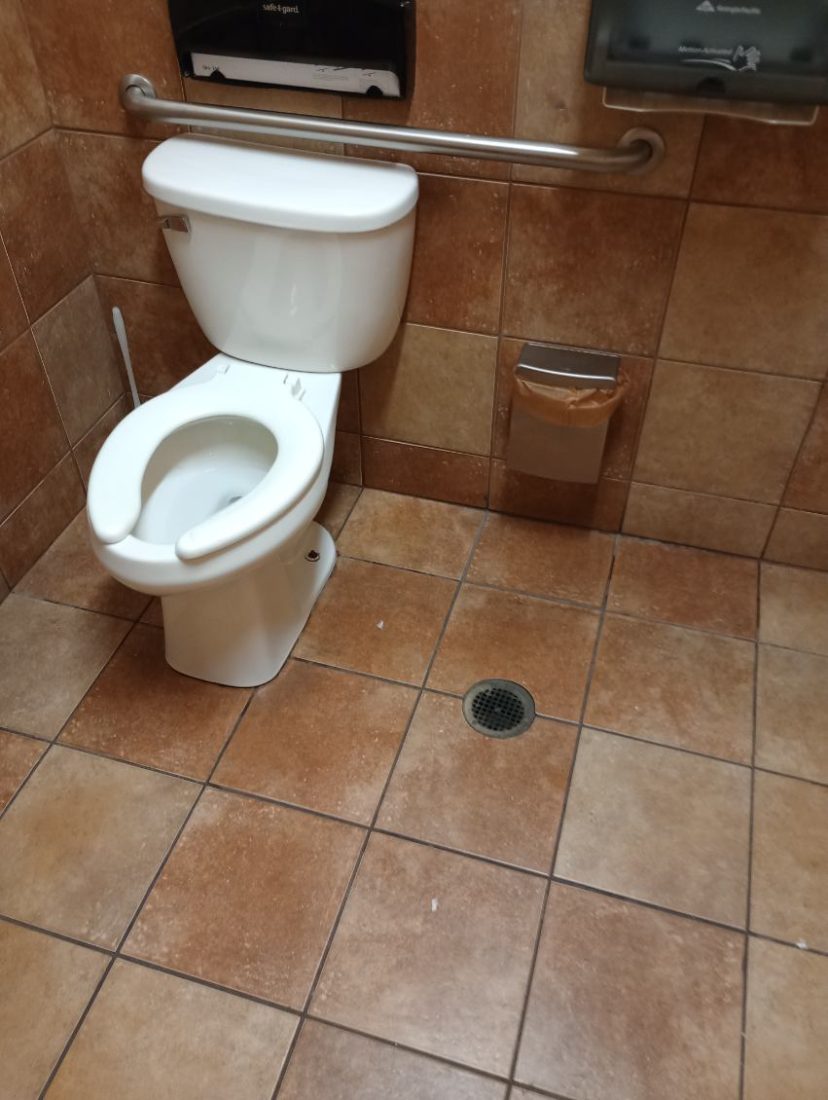 Accessible bathroom stall with white toilet, grab bar, and brown tile flooring