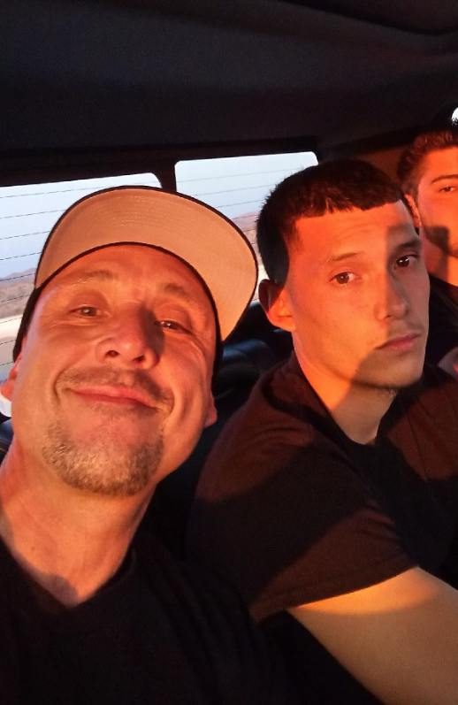 Two men sitting together in a vehicle, one wearing a tan cap and black shirt, the other in dark clothing