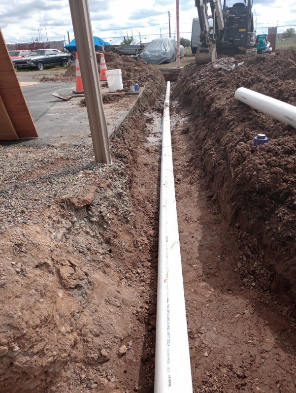 White PVC pipe laid in excavated trench at construction site with heavy equipment