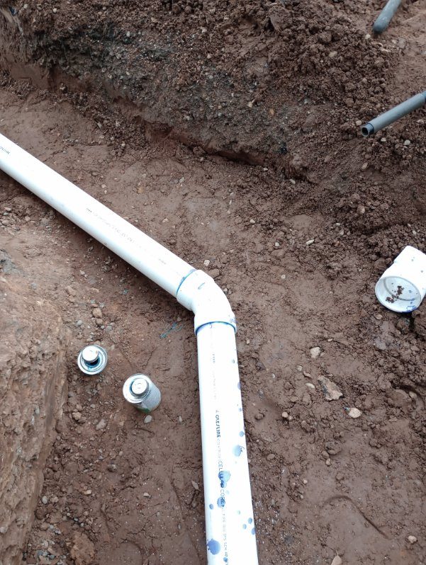 White PVC pipe with elbow connector and metal fittings installed in dirt ground during plumbing construction